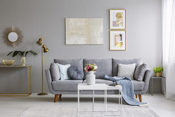 Grey sofa with pillows and blanket standing in bright living room interior with gold lamp, fresh...