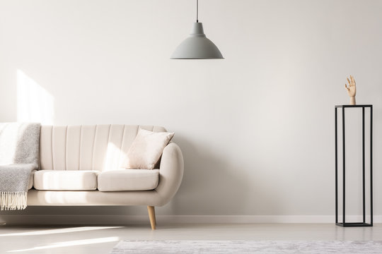 Real photo of a white sofa with cushion and blanket standing next to a black ornament with a wooden hand and a hanging lamp in a simple living room interior