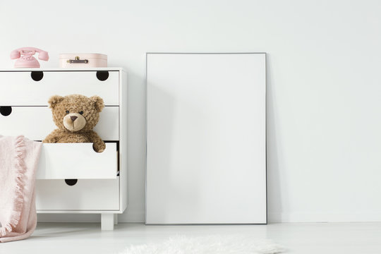 Plush toy and pink blanket in cabinet next to empty white poster in baby's room interior. Real photo. Paste your photo here