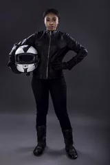 Fotobehang Black female motorcycle biker or race car driver or stuntwoman wearing leather racing suit and holding a protective helmet.  She is standing confidently in a studio © Innovated Captures