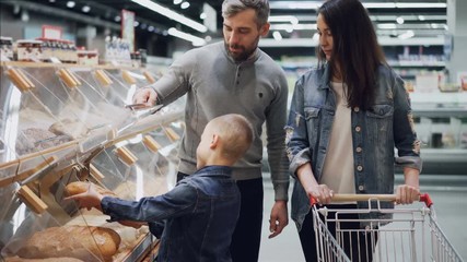 Helpful son is helping his parents to buy bread , father is opening container and boy is taking loaf and smelling it then putting in trolley. Shopping together concept.