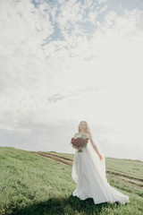 Fototapeta na wymiar Bride with bouquet on sunset outdoor. Boho style. Series. Soft focus. Woman in a beautiful long dress posing in the field. Stunning bride in a wedding dress