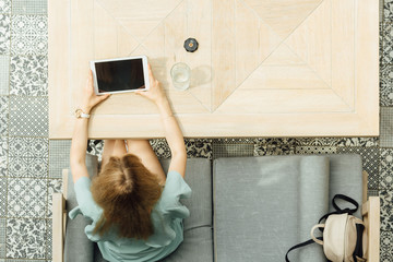 Overhead view of young blond female working with tablet at a cafe. Top view shot of girl sitting at a table with a glass of water browsing internet.