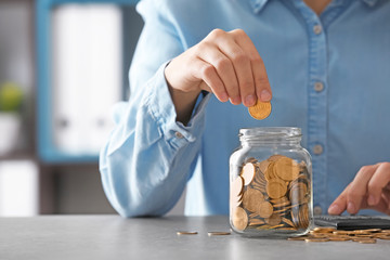 Woman with calculator putting coin into glass jar on table