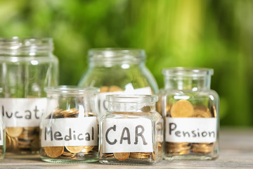 Coins for different needs in glass jars on table outdoors. Money savings concept