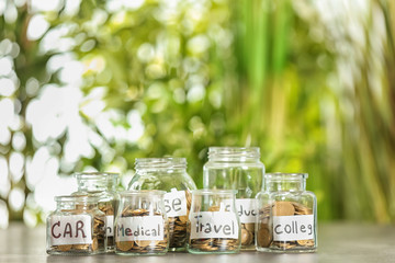Coins for different needs in glass jars on table outdoors. Money savings concept
