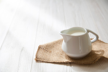 White milk pitcher on a white wooden background. Jug of milk. Farmhouse. Rustic. Sunlit. Copy space.