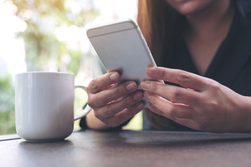 Closeup image of a woman holding and using smartphone with coffee cup in cafe