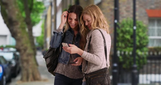 Young female blonde surfs the internet on her mobile device with her friend outside in a nice neighborhood, Two pretty Caucasian women looking at something on a smartphone outdoors, 4k