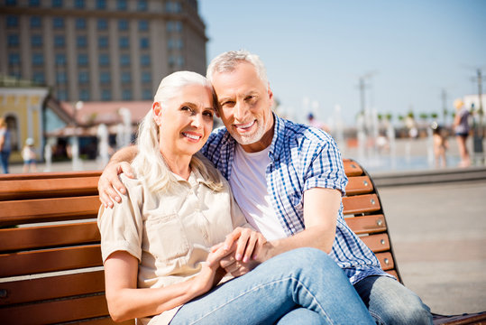 Vacation, weekend, rest, relax, holiday concept! Portrait of lovely romantic grandma and granddad sitting on bench in center of city hugging enjoying good sunny weather
