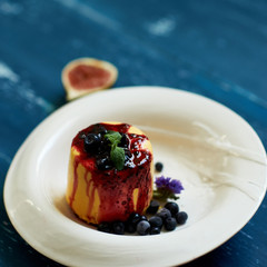 Delicious cake served with blueberries, jam and mint leaves on round plate, fig and cornflowers lying on blue aged wooden surface. Tasty gourmet berry dessert, appetizing sweet meal. Top view.