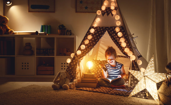 happy child girl laughing and reading book in dark in tent at home