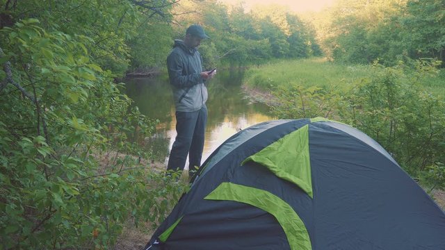 A freelancer man touch a tablet near the forest river and tents.