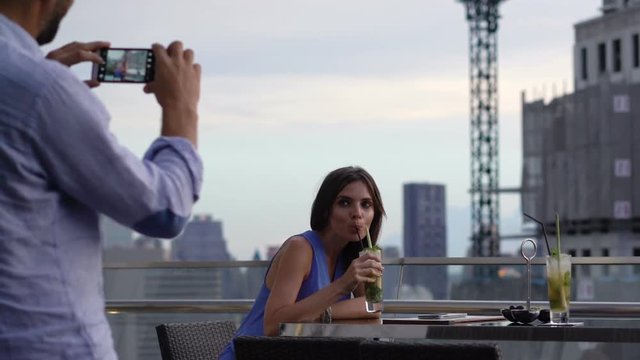 Man taking photo of his beautiful girlfriend sitting on terrace in bar

