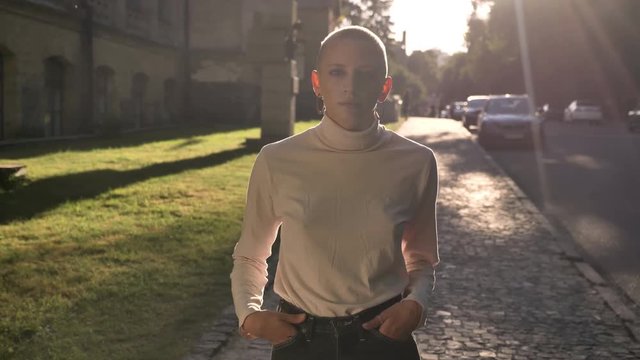 Young confident extraordinary bold girl is standing on street in daytime in summer, puts her hands in pockets, watching at camera