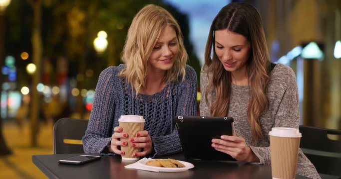 Lovely couple of girls sitting drinking and sharing tablet computer in Champs Elysees, Paris, France, Two attractive young women using portable tablet while drinking at table in European plaza, 4k
