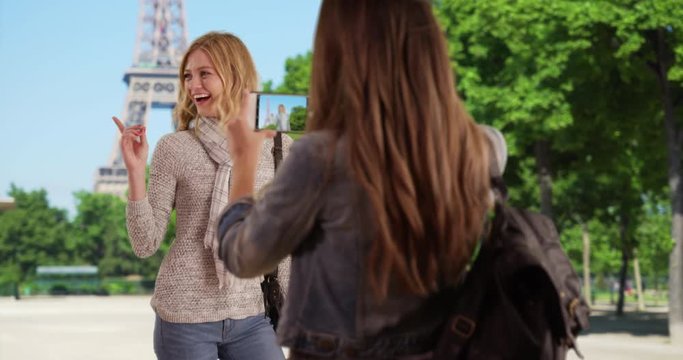 Charming young woman taking picture of friend standing in front of the Eiffel Tower, Attractive caucasian girl using smart phone camera to take girlfriend's picture in Paris, France, 4k