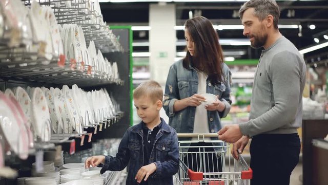 Young family is choosing bowls in supermarket in kitchenware department, they are holding goods comparing them and talking, little boy is helping his parents.