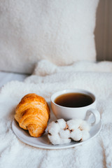 Big white morning cup of coffee and crossant in bed with pillow and cosy blanket decorated with cotton flower.
