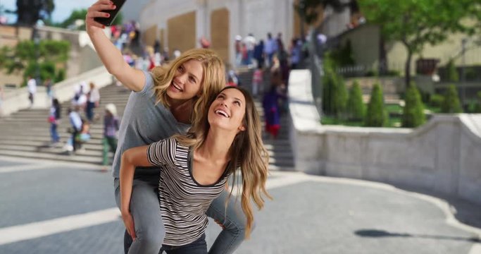 Blonde white girl on her best friend's back taking selfies with her in Rome, Italy, Attractive brunette giving best friend piggyback ride while she takes selfies on phone near the Spanish Steps, 4k