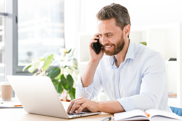 Handsome bearded man talking by mobile phone using laptop computer.