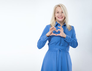 Friendly smiling middle aged woman in blue dress looks at the camera and shows a gesture I love you isolated on white.