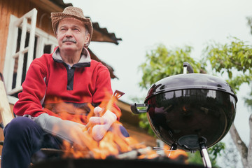 Barbecue time. European man in hat waiting for grilled food. He is sitting near fire and rest.