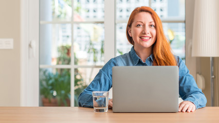 Redhead woman using computer laptop at home with a happy face standing and smiling with a confident smile showing teeth