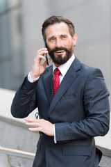 Business man talking active with the client on a cell phone out of office building. Handsome bearded manager outdoor talking on the phone at business center in the background.