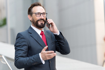 A good deal! Bearded businessman speaks by phone and laughs.View of a Young attractive business man in glasses using smartphone. Smiling formal dressed businessman talking on cell phone with customers
