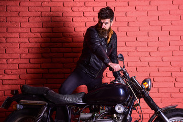 Fototapeta na wymiar Masculine passion concept. Man with beard, biker in leather jacket near motor bike in garage, brick wall background. Hipster, brutal biker on serious face in leather jacket gets on motorcycle.
