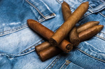 Pile of thick cigars, denim background. Cigars lay on back pocket of blue jeans. Collection of elite cigars. Habits concept. Habit to smoke elite tobacco