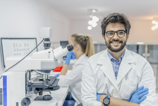 Closeup portrait, young friendly scientist standing by microscope. Isolated lab background. Research and development sector