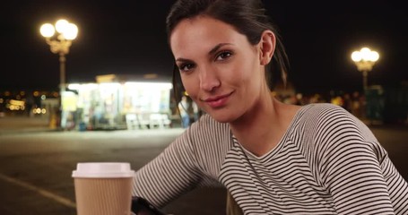 Portrait of millennial woman sitting with coffee by gas station at night, Caucasian girl wearing striped shirt seated with coffee cup outdoors near petrol station, 4k