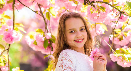 Fototapeta premium Cute child enjoy aroma of sakura on spring day. Girl on smiling face standing near sakura flowers, defocused. Perfume and fragrance concept. Girl with long hair outdoor, cherry blossom on background.