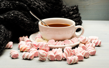 Mug and sweets near cozy knitted scarf. Mug filled with black brewed tea, spoon and heap of marshmallow on white wooden background. Coziness concept. Cup with hot black tea and marshmallows
