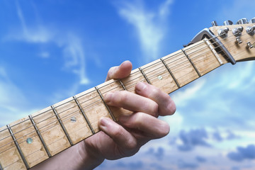 Guitarist's hand playing and fretting guitar strings under a blue sky.