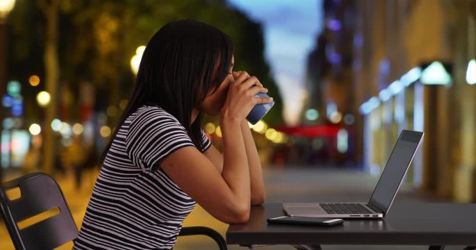Pretty black woman on city street at night drinking coffee and browsing on laptop, Side view of business woman sitting outdoors using her computer, 4k