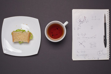A bacon and green sandwich on a white plate. A Cup of tea next to a plate of sandwiches. Student's Breakfast. A sheet of paper with formulas and numbers on the table.