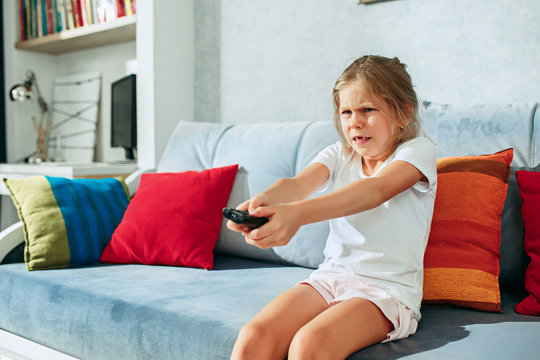 Little casual girl watching tv at home. Female kid sitting on sofa with TV remote and switching channels