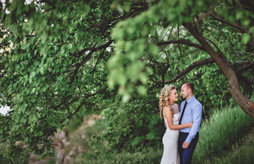 Beautiful newlyweds stand in a green grove, near a tree, hug and smile. A stylish bridegroom embraces a sweet bride with curly hair at a wedding.