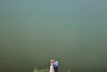 Fototapeta premium Stylish newlyweds stand on the background of the lake. A young groom and a beautiful bride with curly hair stand against the sea and embrace. Wedding.