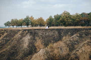 A stylish bridegroom in a black suit and a beautiful bride in a lace dress stand against the background of gray rocks, hills and blue clouds. Autumn wedding portrait of cute newlyweds. Film effect.