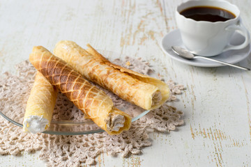 Sweet dessert. Waffle tubules with a delicate protein cream on a transparent round plate, next to a cup of coffee on an old wooden table.