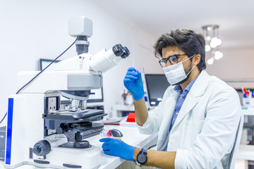 Scientist in lab coat and sterile mask doing microscope analysis.