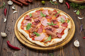 Delicious italian pizza with ham, bavarian sausages and prosciutto decorated by basil leaves on wooden background