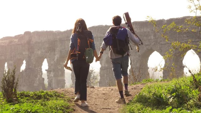 Young lovely couple backpackers tourists walking holding hands toward roman aqueduct arches in parco degli acquedotti park ruins in rome on romantic misty sunrise with guitar and sleeping bag slow