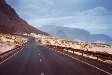 Asphalt road leads between black volcano erosion mountain and white sand dunes. Breathtaking coastline of Calhau, Sao Vicente Island Cape Verde