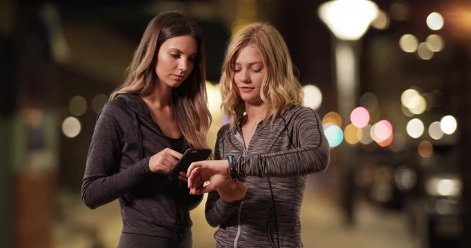 Pair of pretty young ladies in workout attire checking smartphone and wristwatch outside at night, Couple of attractive fitness girls standing outside at night using phone and watch, 4k