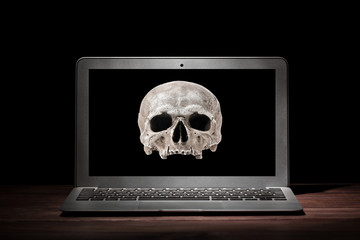 Cybercrime or hacking concept. Laptop in dark room under beam of light with a skull on a display. Idea of virus or worm program cyber attack.